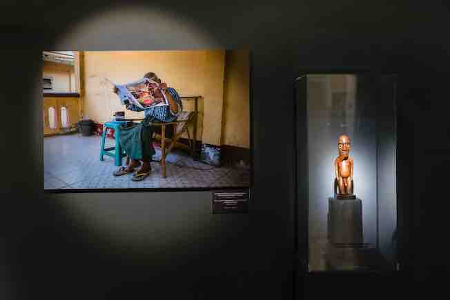 Photographie de Steve McCurry montrant un homme lisant le journal dans une maison pour personnes âgées à Mingun, au Myannmar, 2017, associée à une statuette moai miro de l’île de Pâques conservée au musée Barbier-Mueller. Photo Luis Lourenço.